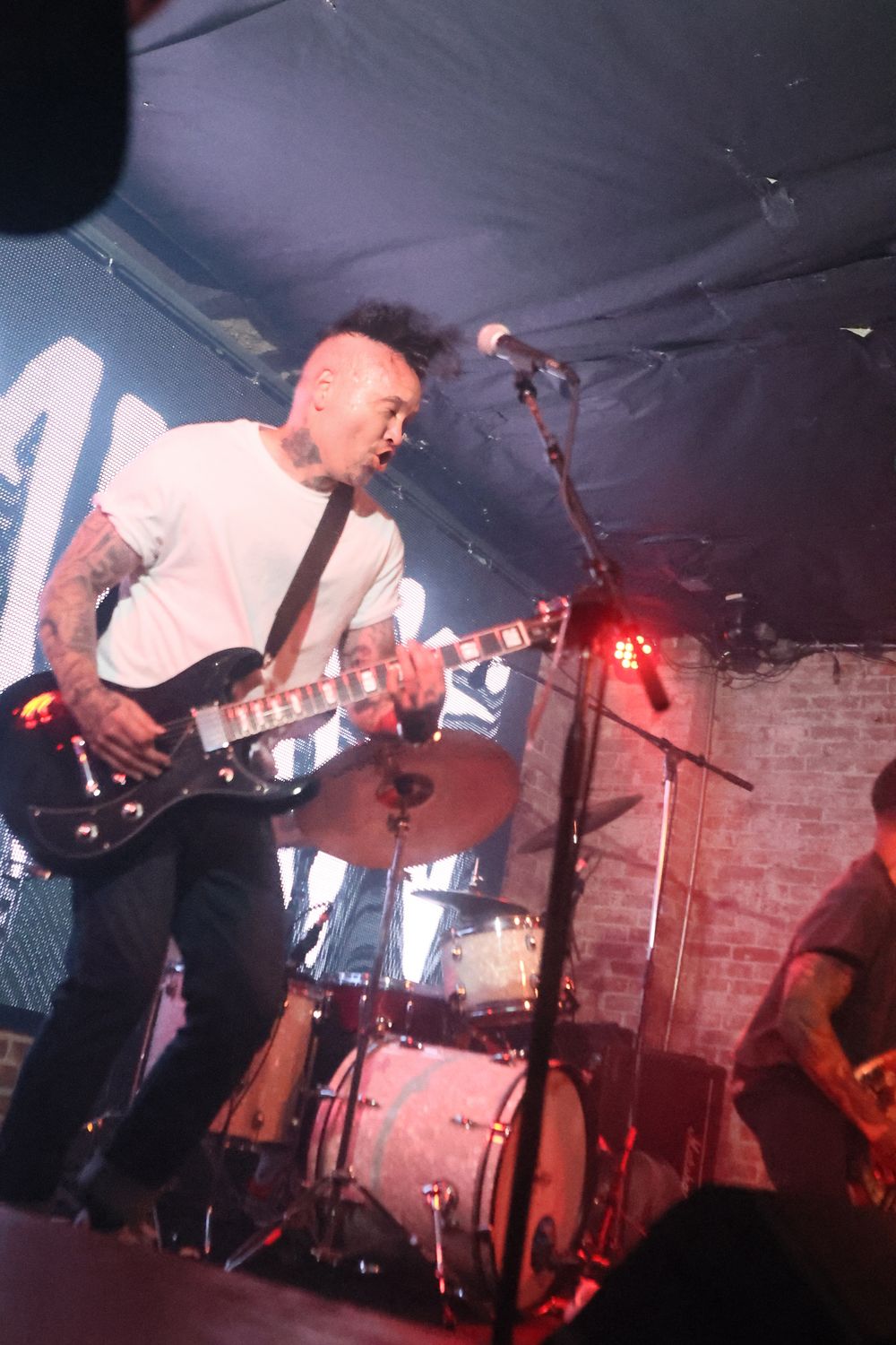 Musicians perform energetically on a dimly lit stage with red lighting and exposed brick walls in the background.