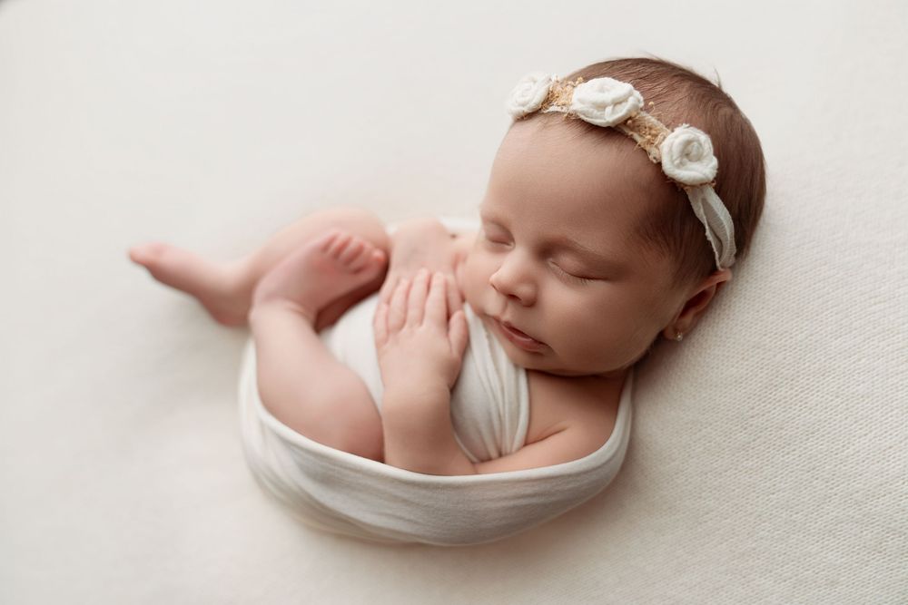 Newborn baby girl sleeping peacefully wrapped in white, wearing a floral headband, in a professional studio photo.