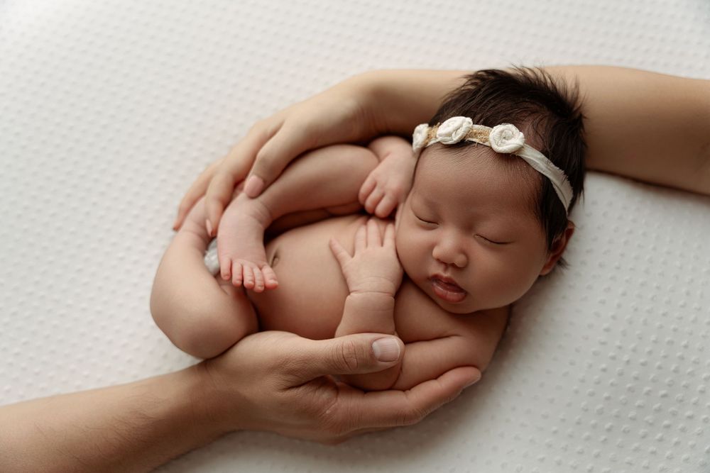 Newborn baby sleeping peacefully in parents hands, wearing white floral headband, on white background.