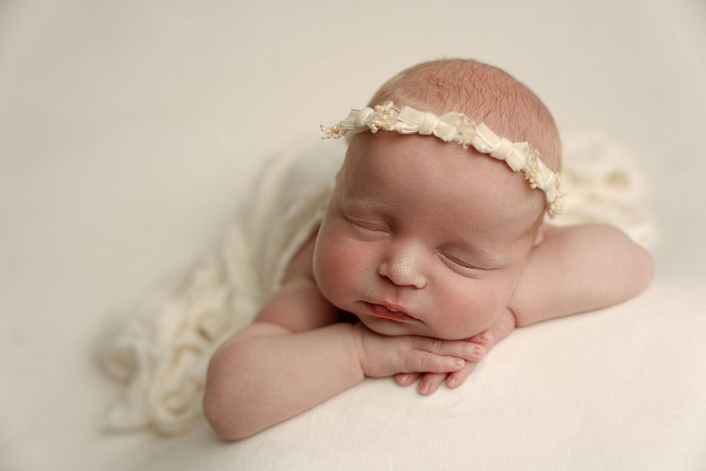 Sleeping newborn baby girl wearing a delicate white floral headband, posed with hands under chin on a soft cream blanket.