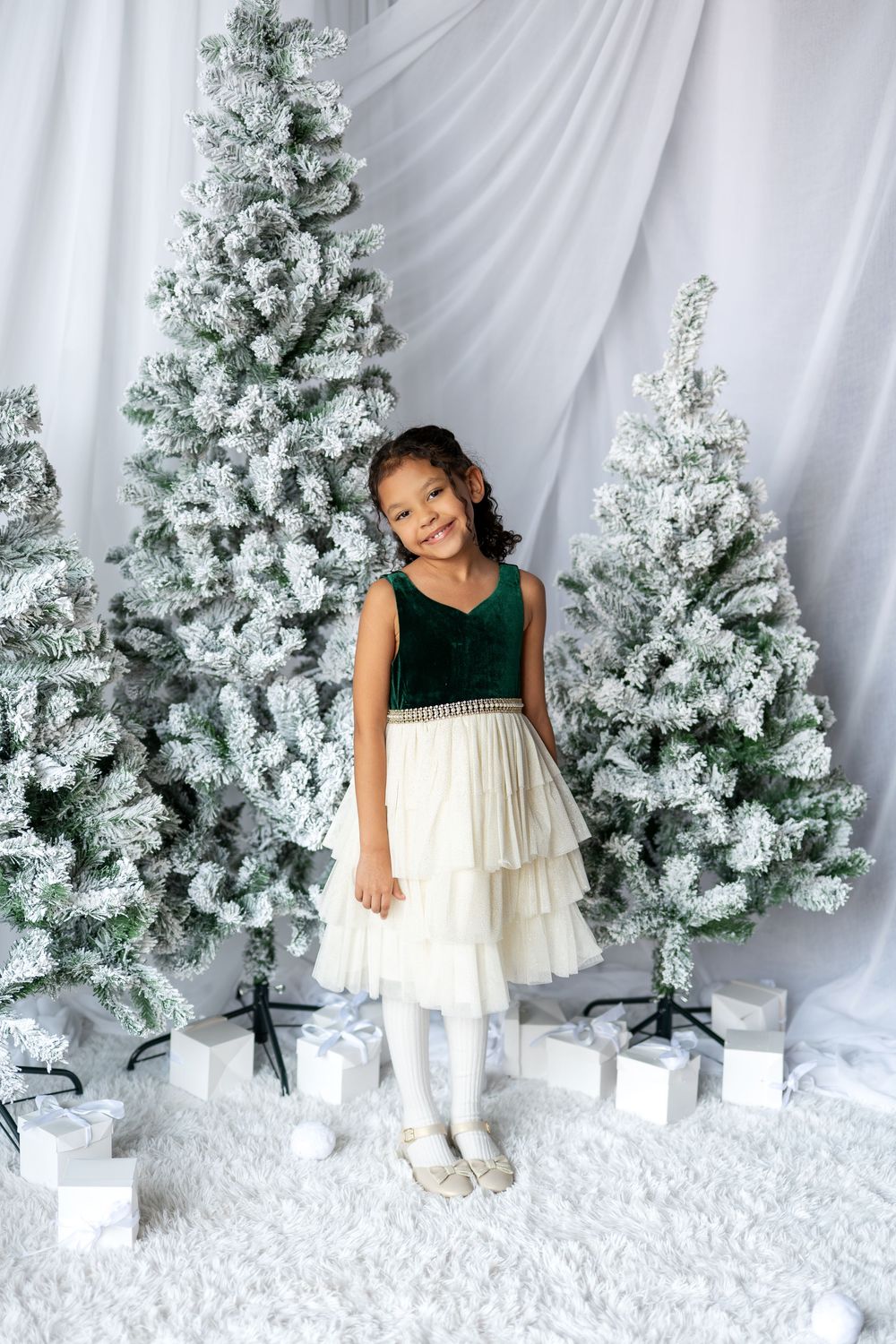 Young girl in green and white holiday dress standing among snow-flocked Christmas trees with gifts.