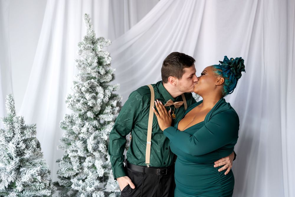 Interracial couple sharing a kiss in matching green outfits during a festive Christmas photo session with snowy trees.