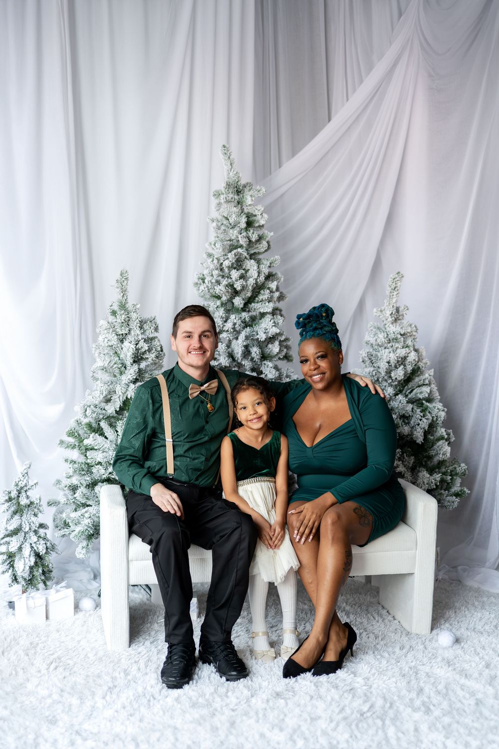 Interracial family of three posing on white bench surrounded by snow-flocked Christmas trees in coordinating green outfits.