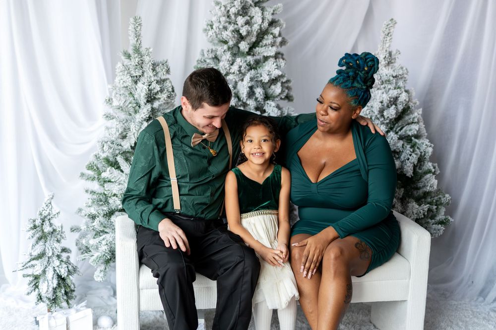 Loving interracial family sharing a tender moment on white bench with snow-covered Christmas trees in background.
