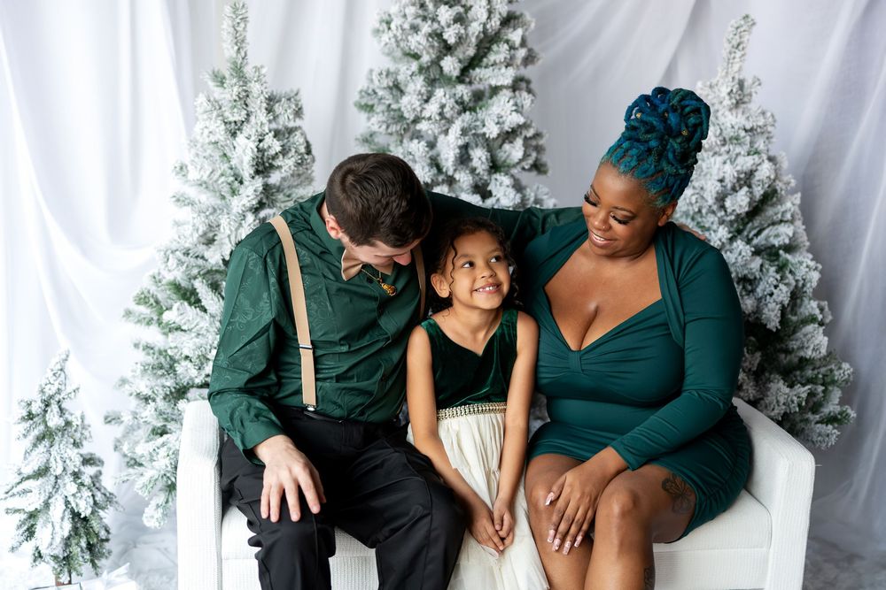 Interracial family of three sharing a warm embrace on white bench surrounded by flocked Christmas trees.