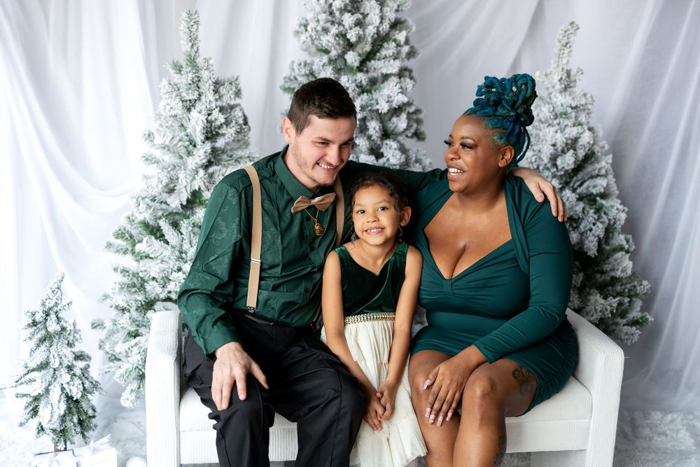 Happy interracial family of three smiling together on white bench with snow-flocked Christmas trees behind them.