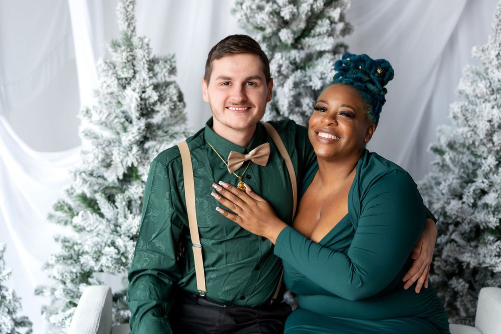 Smiling interracial couple in matching emerald green outfits posing together before snow-flocked Christmas trees.