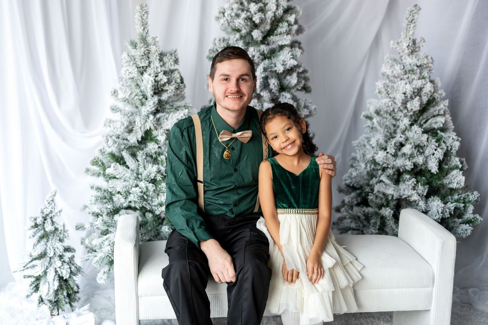 Father and daughter in coordinating outfits smiling together on white bench with snow-flocked Christmas trees.
