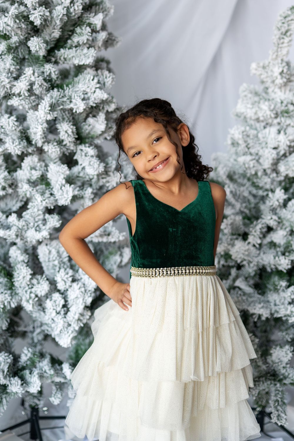Young girl smiling in green velvet and white tulle dress with Christmas trees behind her.
