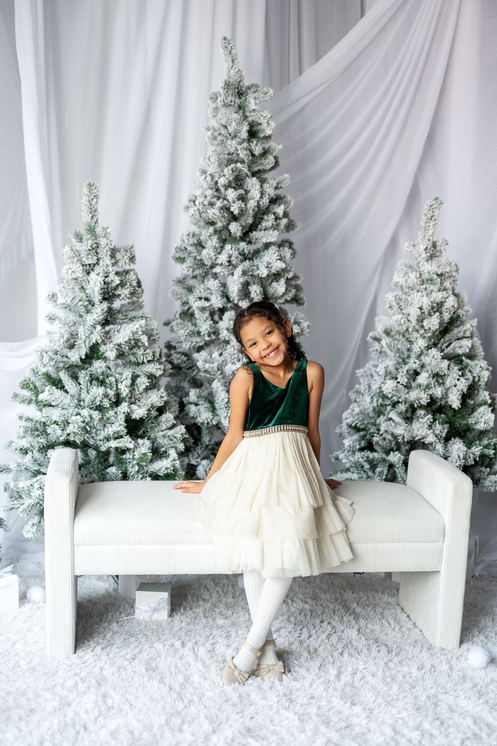 A young girl in a green velvet and cream tulle dress sits on a white bench surrounded by snow-flocked Christmas trees.