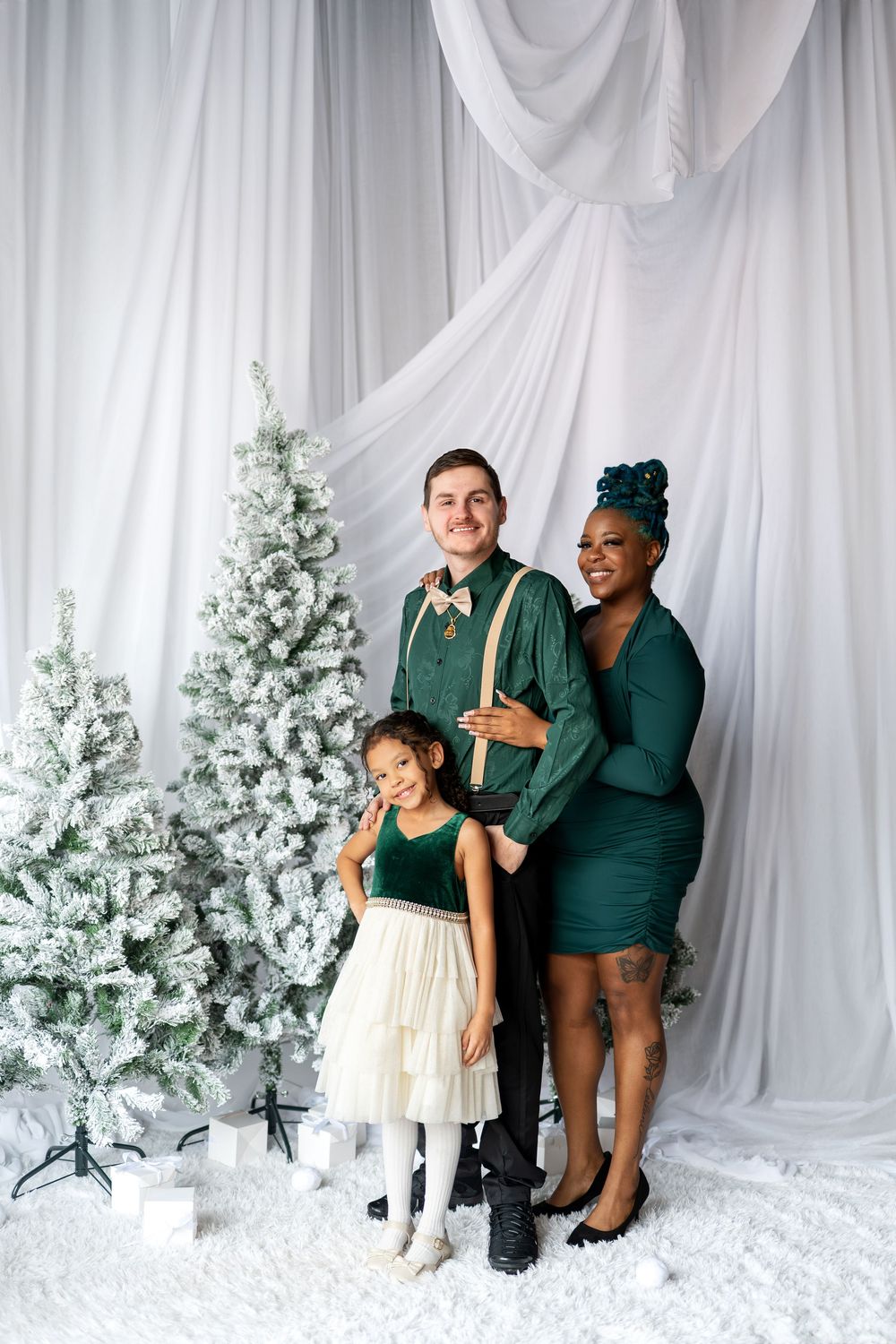A mixed-race family of three in coordinating green outfits poses together beside snow-flocked Christmas trees.