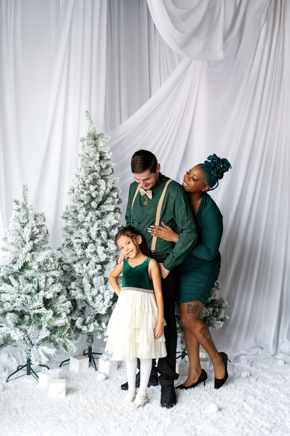 A joyful mixed-race family dressed in green holiday outfits laughs together near snow-flocked Christmas trees.