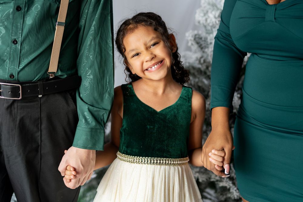 A happy young girl in a green velvet dress holds hands with her parents during a Christmas photo session.