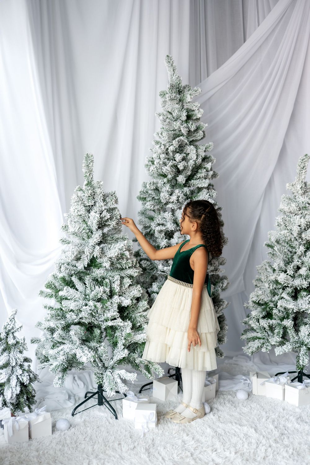 A young girl in a green velvet dress reaches up to touch a snow-flocked Christmas tree in a white studio setting.