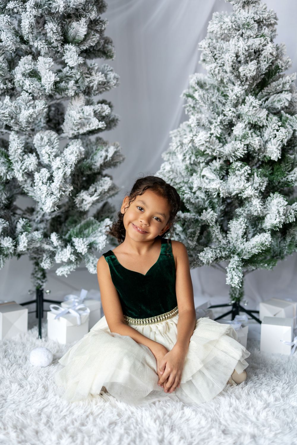Smiling young girl in green velvet dress sitting on white fur rug near Christmas trees and gifts.