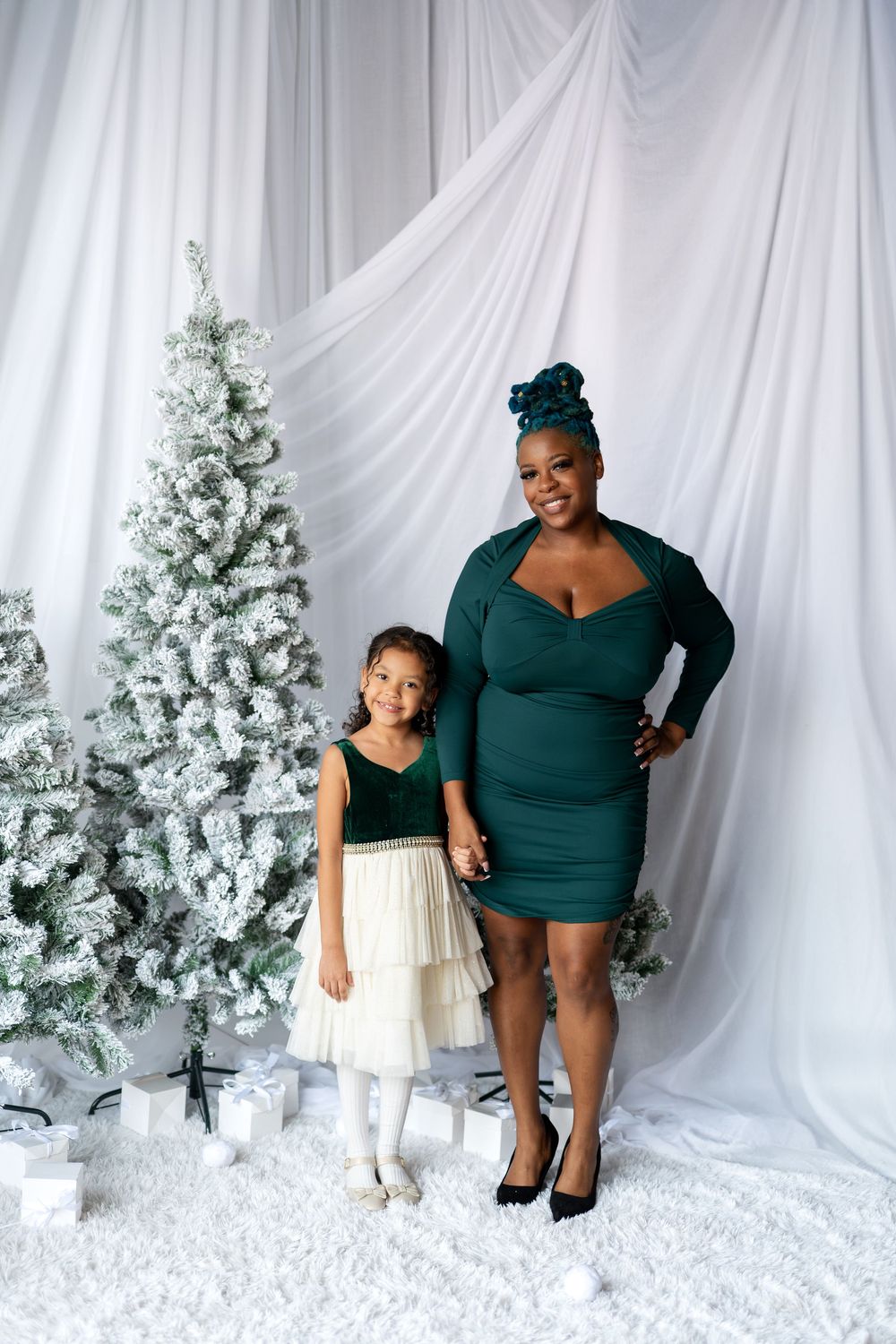 Mother and daughter in matching green outfits holding hands by flocked Christmas tree with gifts.