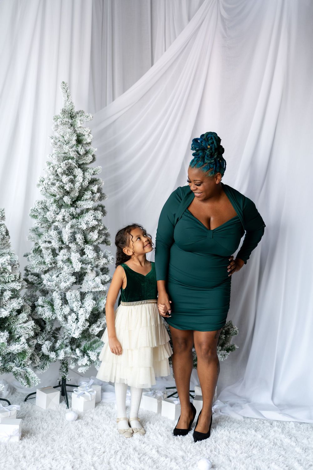 Mother in green dress looking down at daughter in holiday dress near snow-flocked Christmas trees.