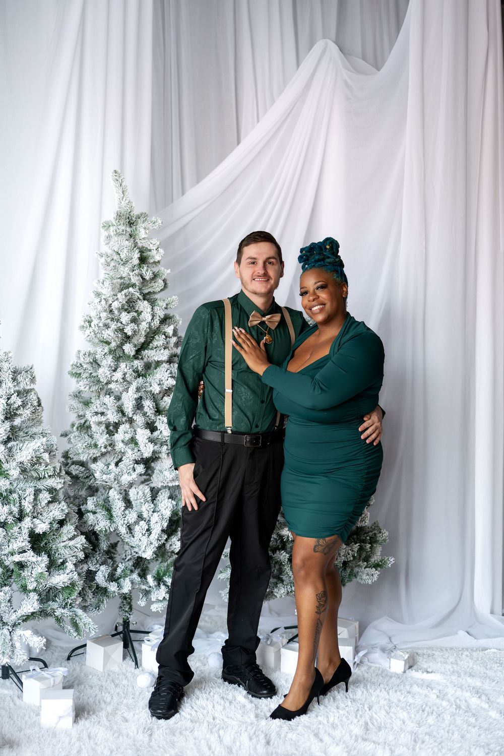 Interracial couple in coordinating green holiday outfits posing by snow-flocked Christmas trees.