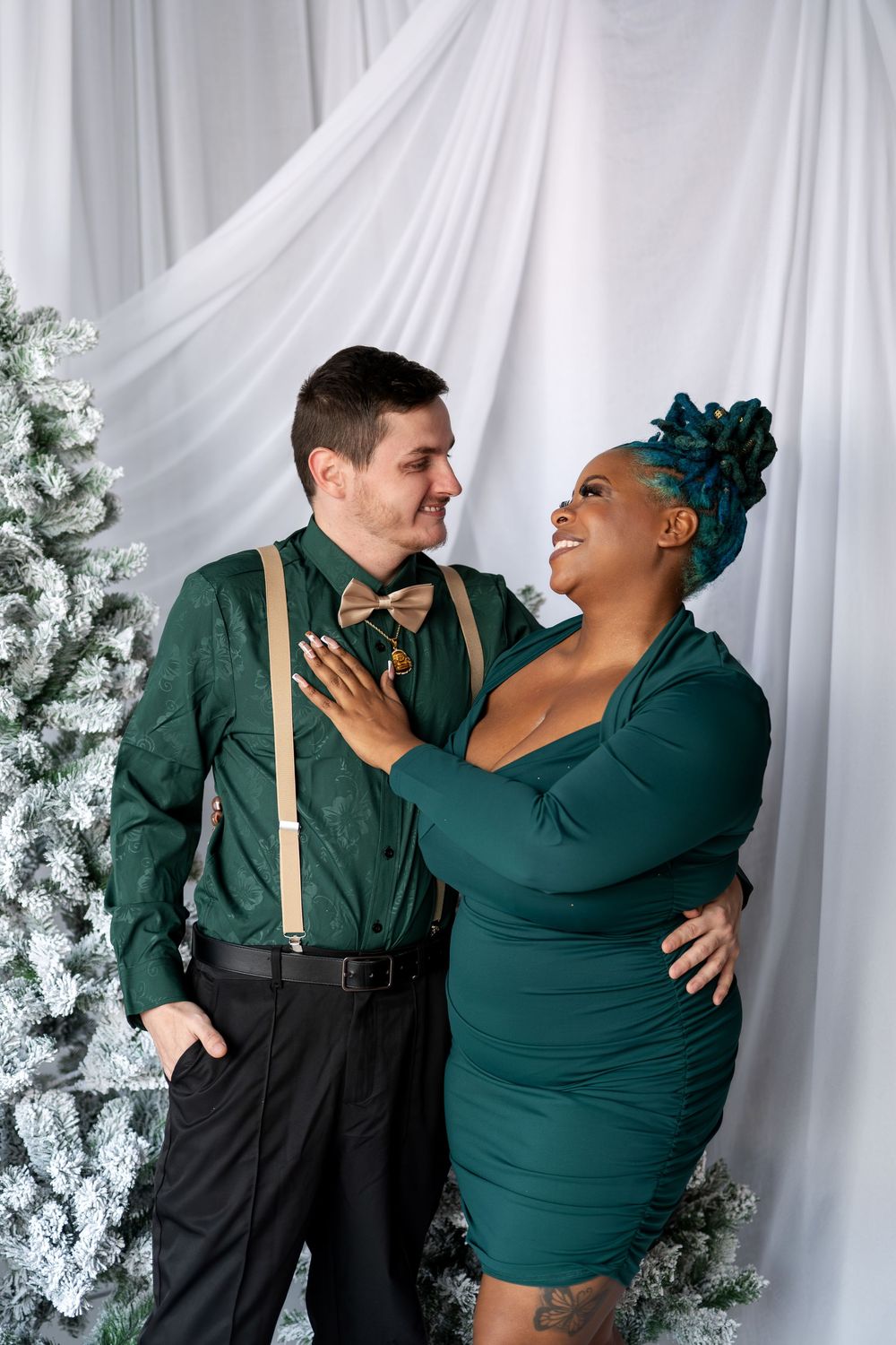 Smiling interracial couple in green outfits gazing at each other by flocked Christmas trees.