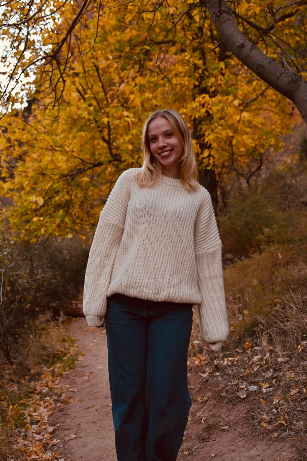 Smiling blonde woman in cream knit sweater walks along autumn forest trail with golden leaves.
