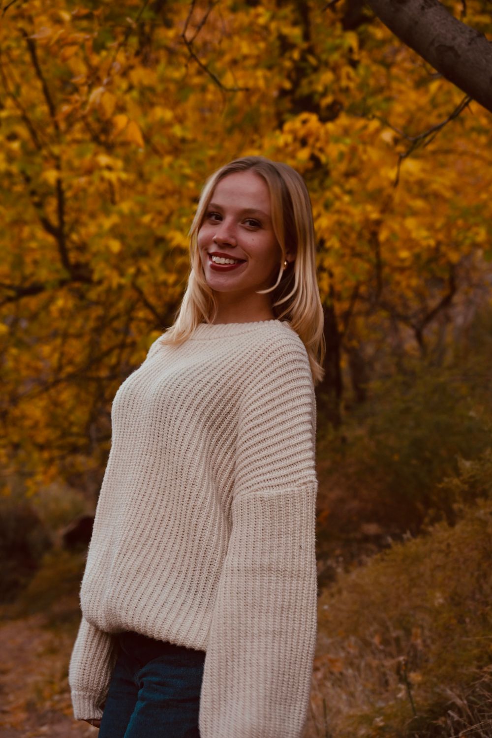 Blonde woman in cream ribbed knit sweater smiles against vibrant yellow autumn foliage backdrop.