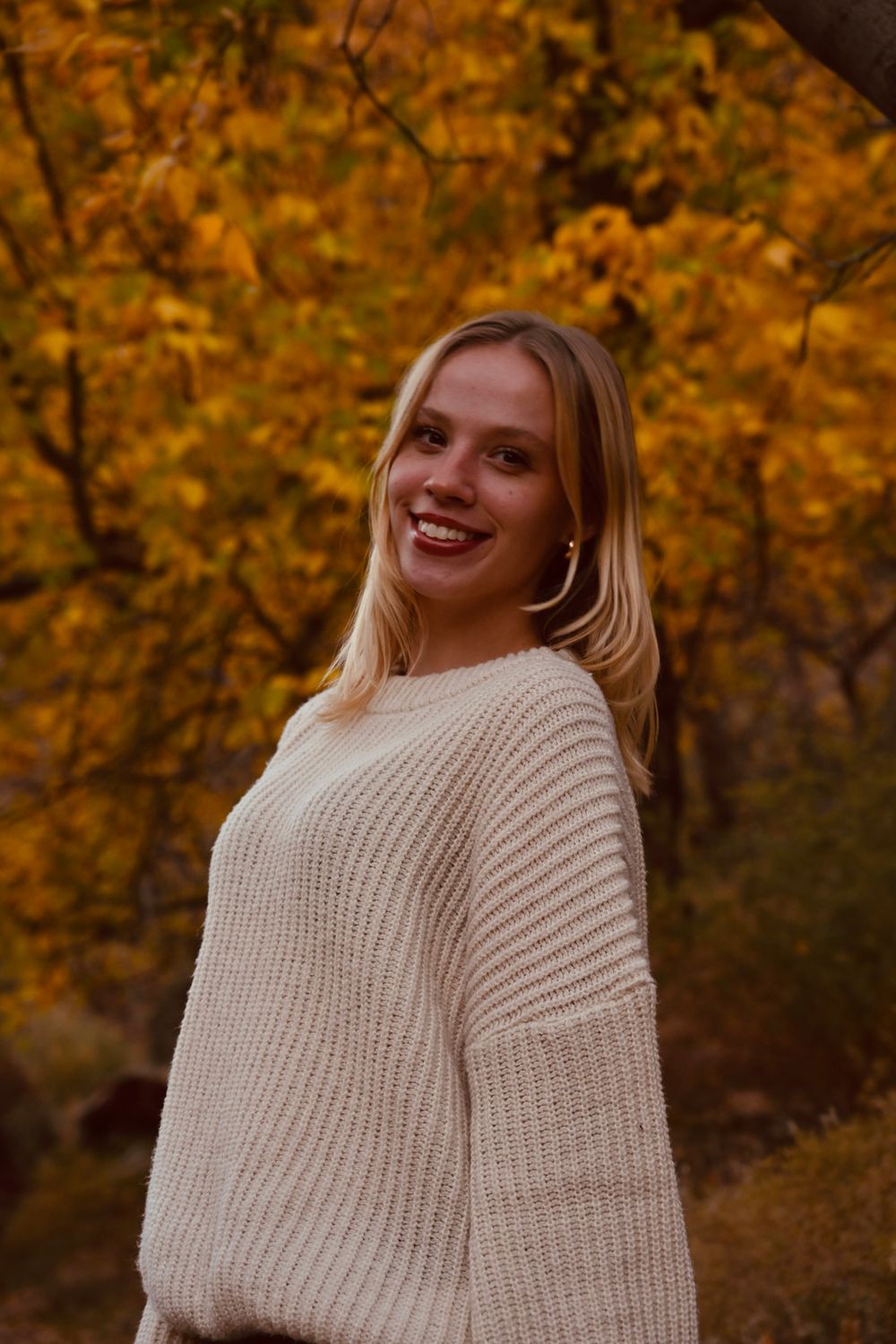 Smiling blonde woman in cream knit sweater poses among golden autumn trees in fall setting.