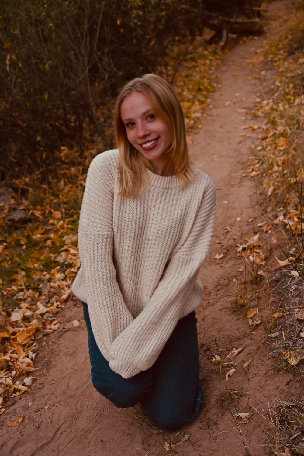 Young blonde woman kneeling on autumn trail wearing cream knit sweater and jeans surrounded by fall foliage.