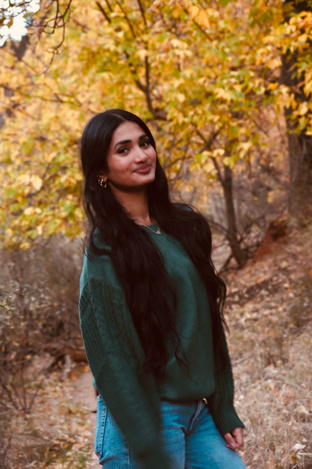 Young woman with long black hair wearing green sweater and jeans posing among golden autumn trees.