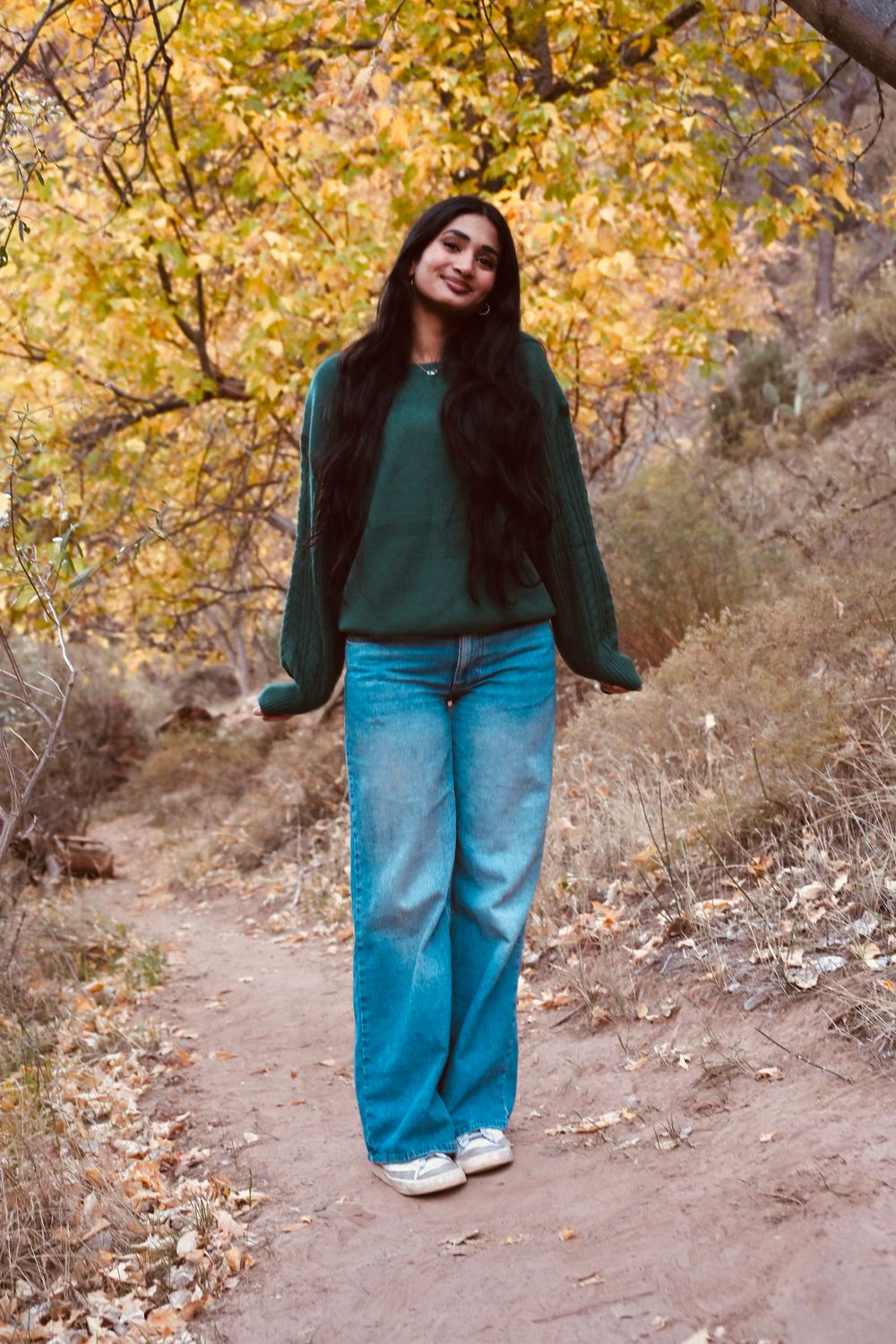 Young woman in green sweater and wide-leg jeans walking on a dirt trail surrounded by autumn trees.