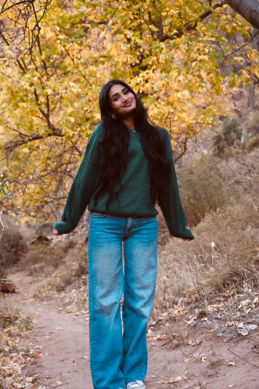 Smiling young woman with long dark hair in green sweater and jeans strolling along autumn nature path.