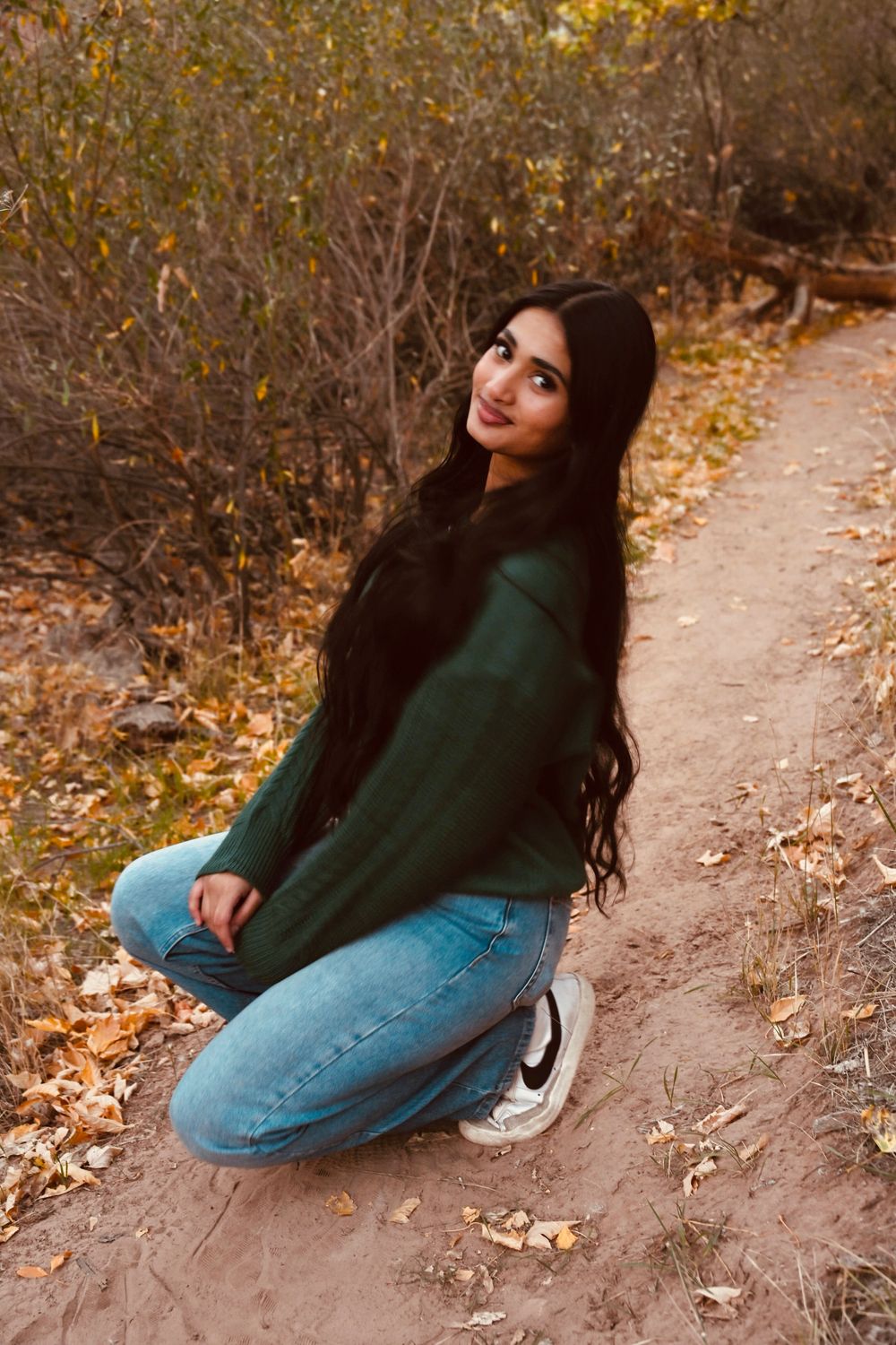 Young woman with long black hair in green sweater and jeans sitting on sandy trail in autumn forest.