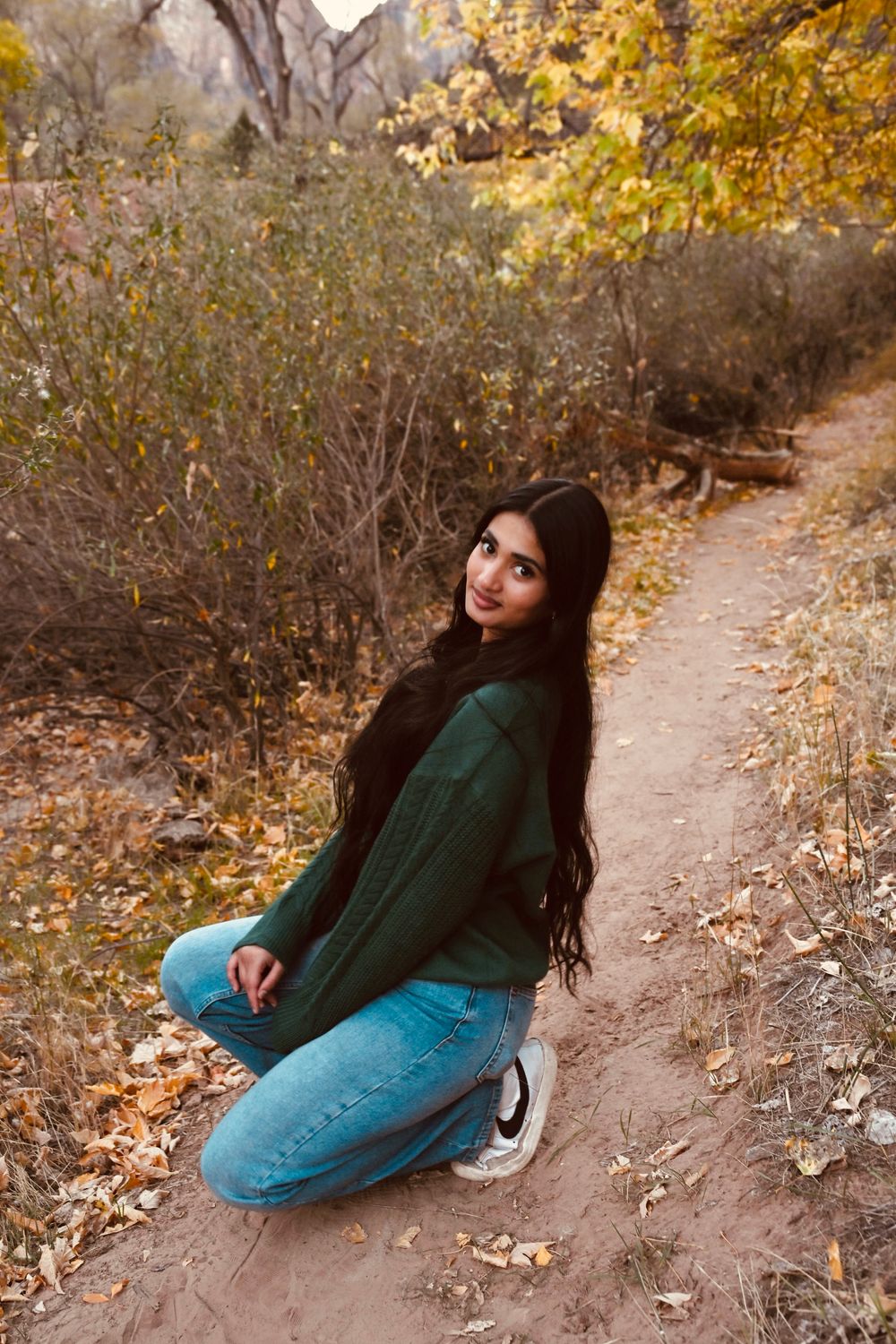 Young woman in green sweater and jeans sitting on dirt path surrounded by golden fall foliage and rocks.