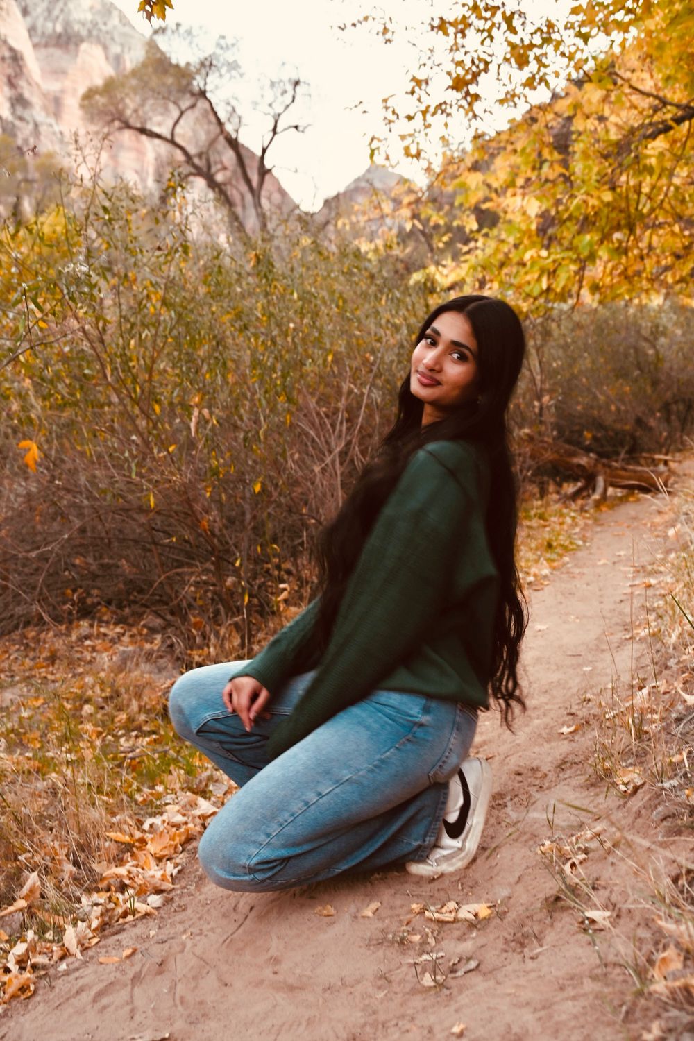 Young woman in green sweater and jeans kneeling on autumn trail with red canyon cliffs in background.