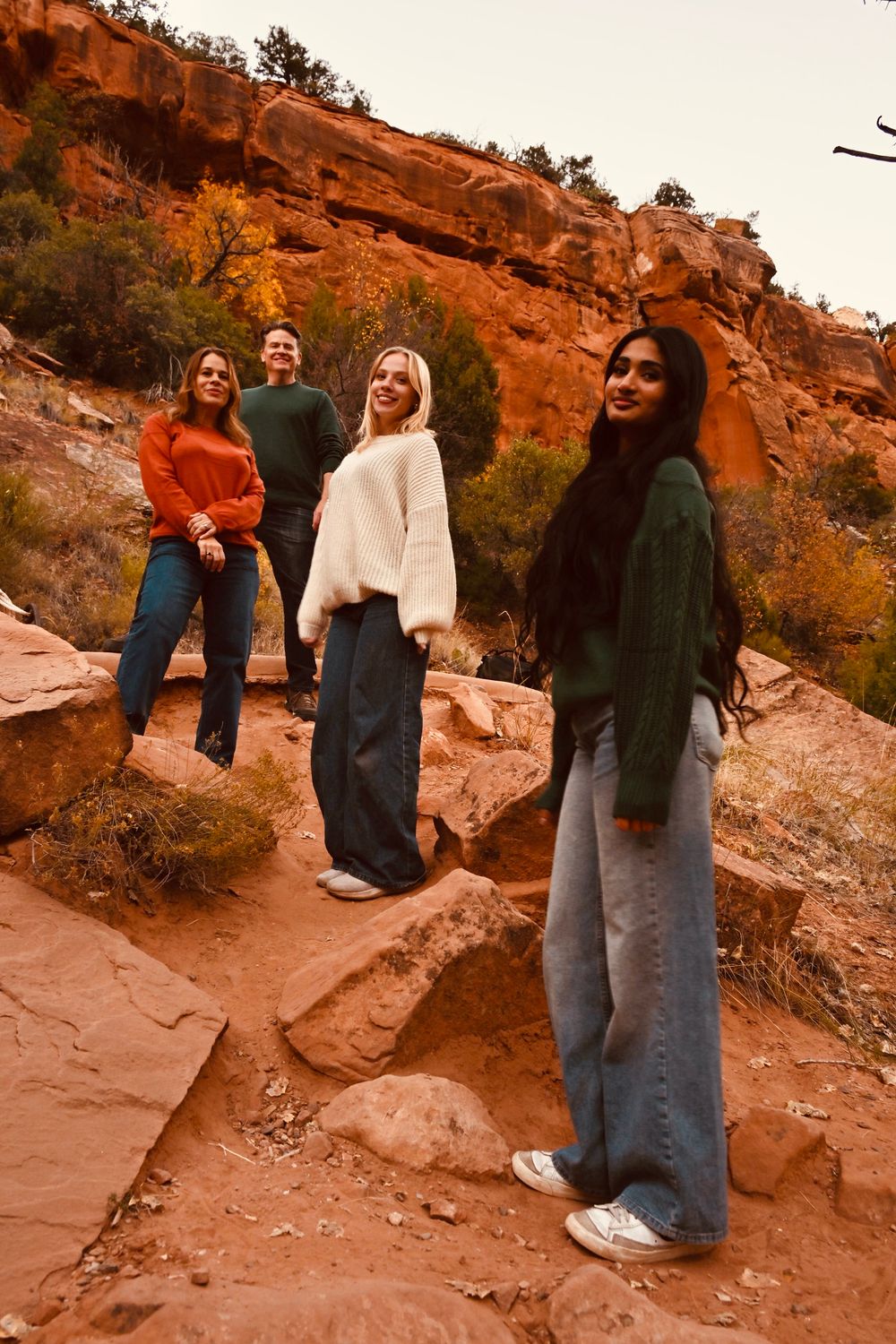 Group of four friends posing on rocky red canyon trail wearing colorful fall sweaters and jeans.