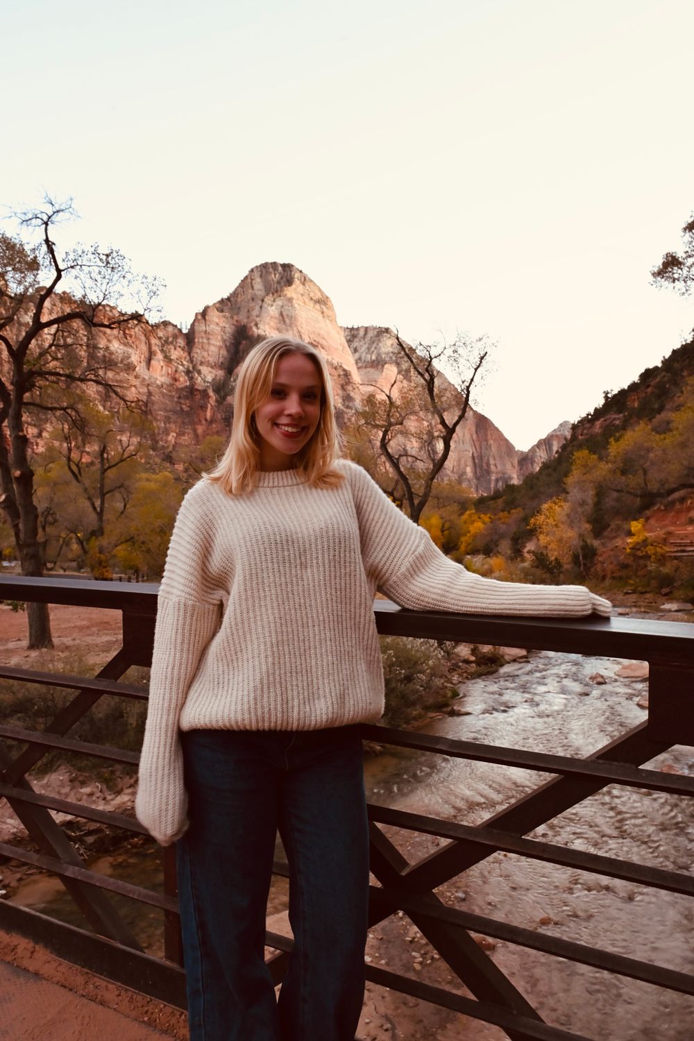 Blonde woman in cream knit sweater smiling on bridge at Zion National Park with river and red rock mountains.