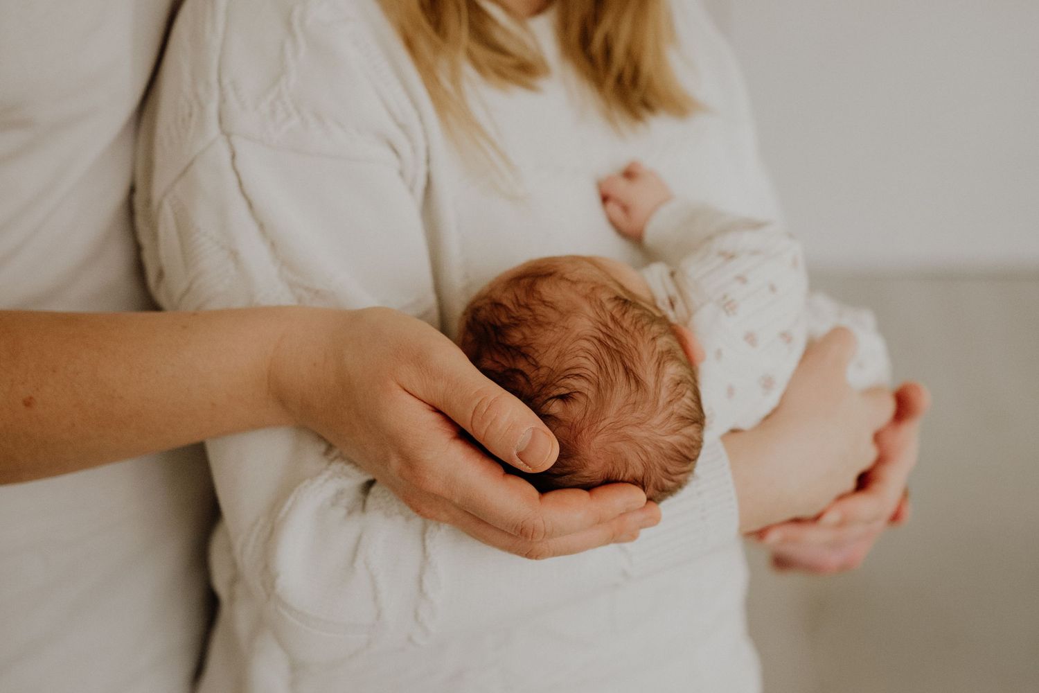 Baby in white onesie being held by adult hands against neutral background.