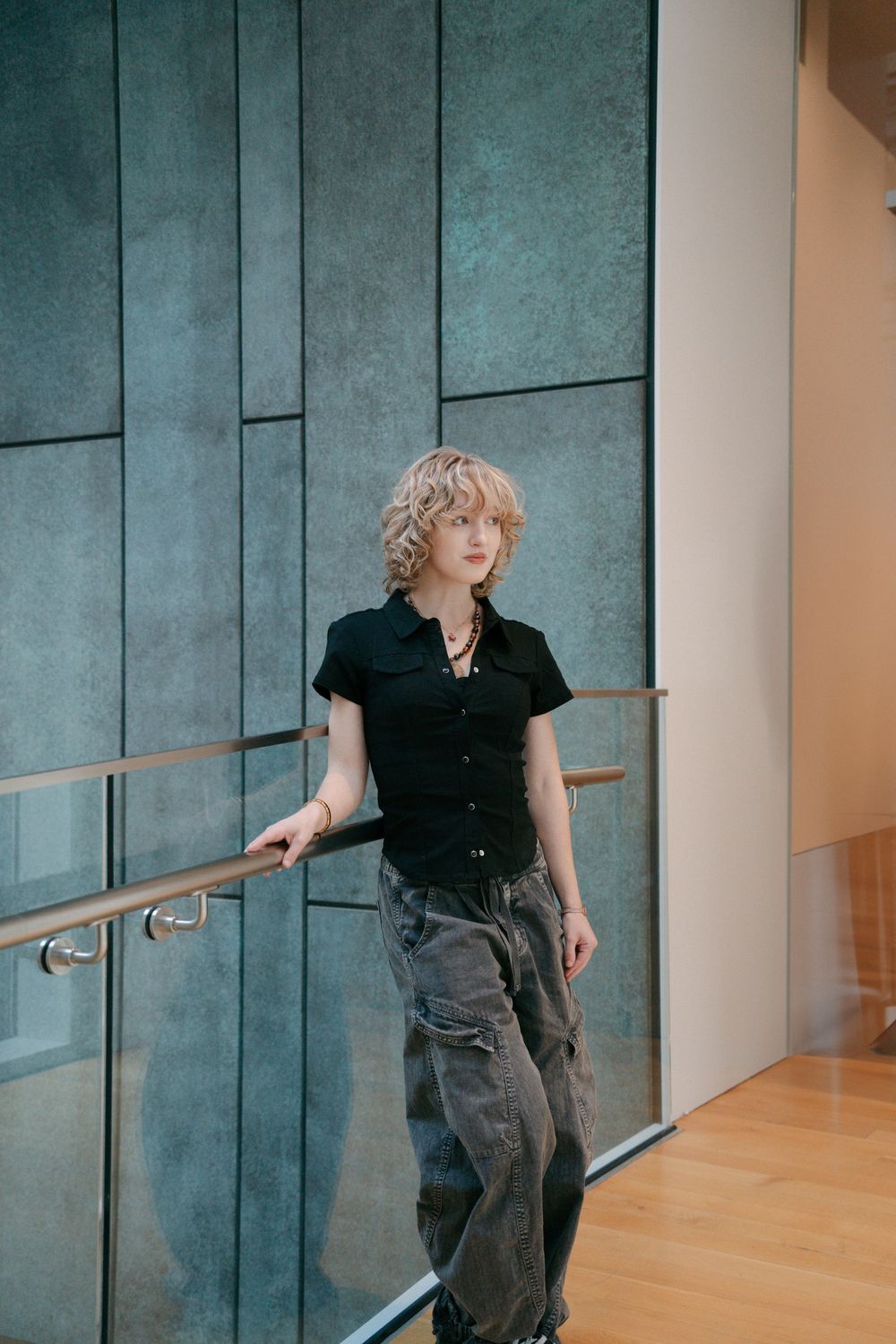 Blonde woman in black shirt and cargo pants leaning on railing indoors.