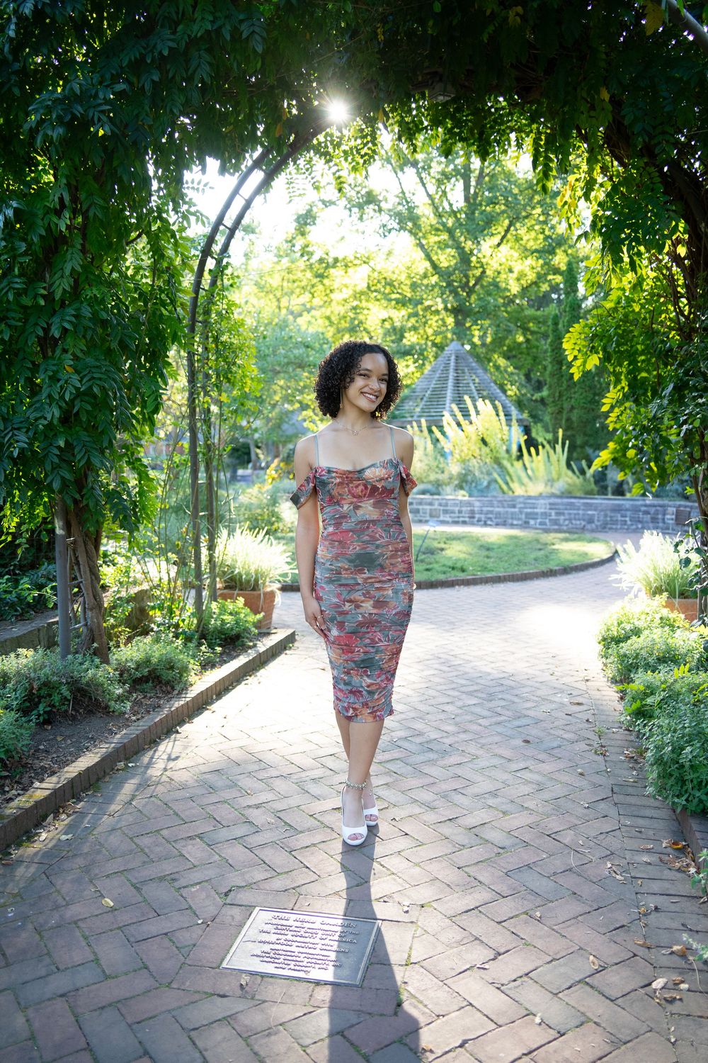 Woman in floral midi dress walking through lush garden archway in sunlight.