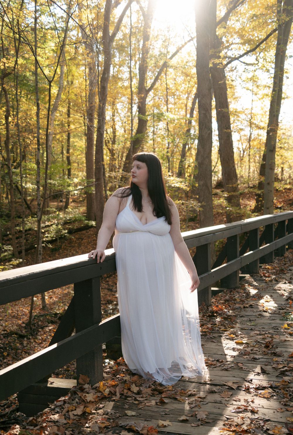 Woman in white gown standing on wooden bridge in golden autumn forest.