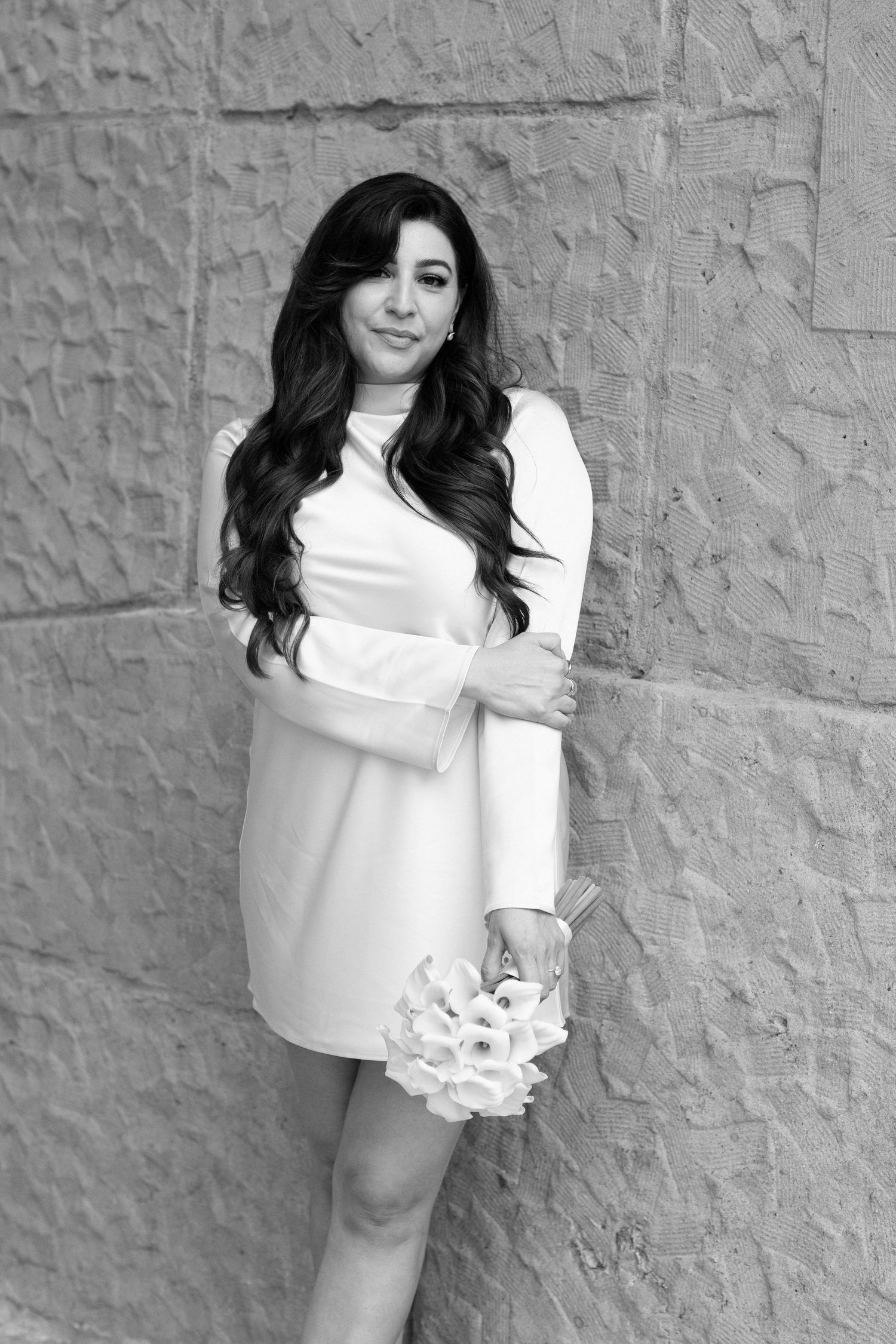 Black and white portrait of a person in a white long-sleeved dress standing against a textured wall.