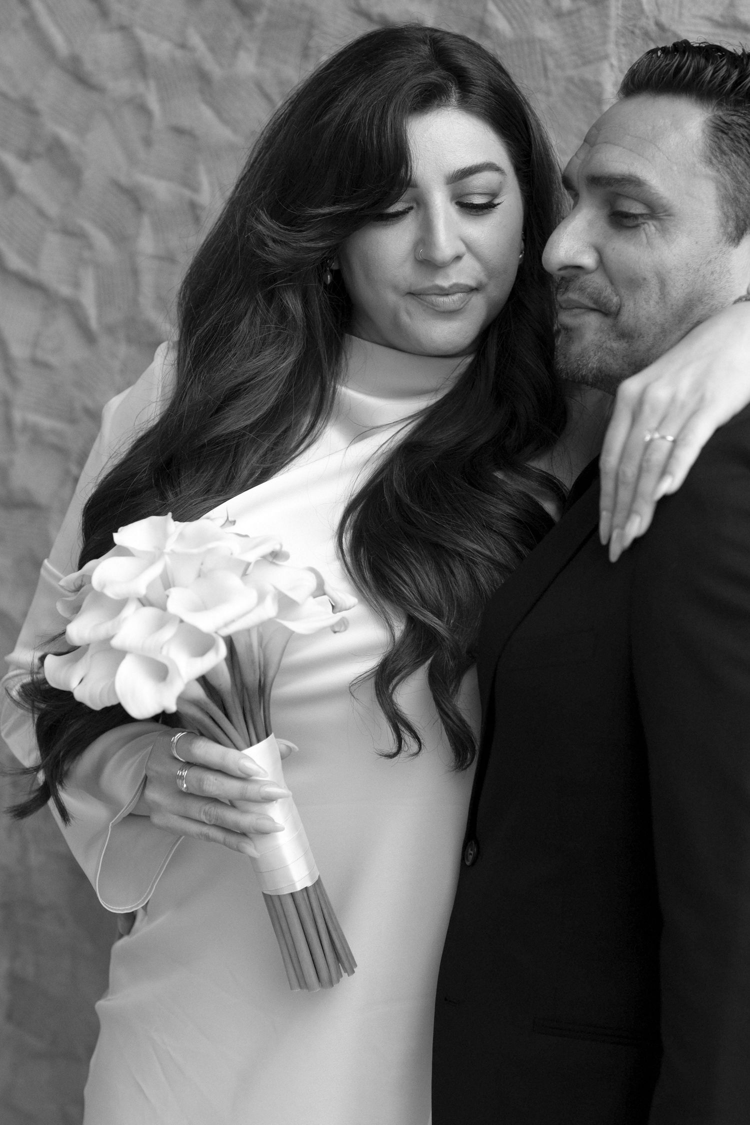 A romantic black and white wedding portrait showing a couple embracing while holding a white calla lily bouquet.