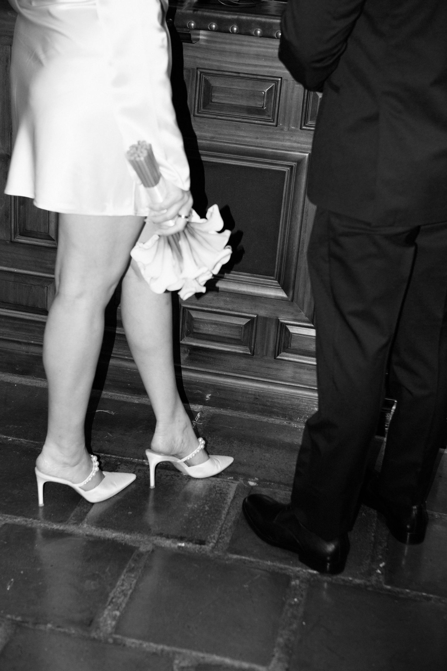 Black and white photo showing white heels and a dress hem next to dark suit pants standing on tiled floor.
