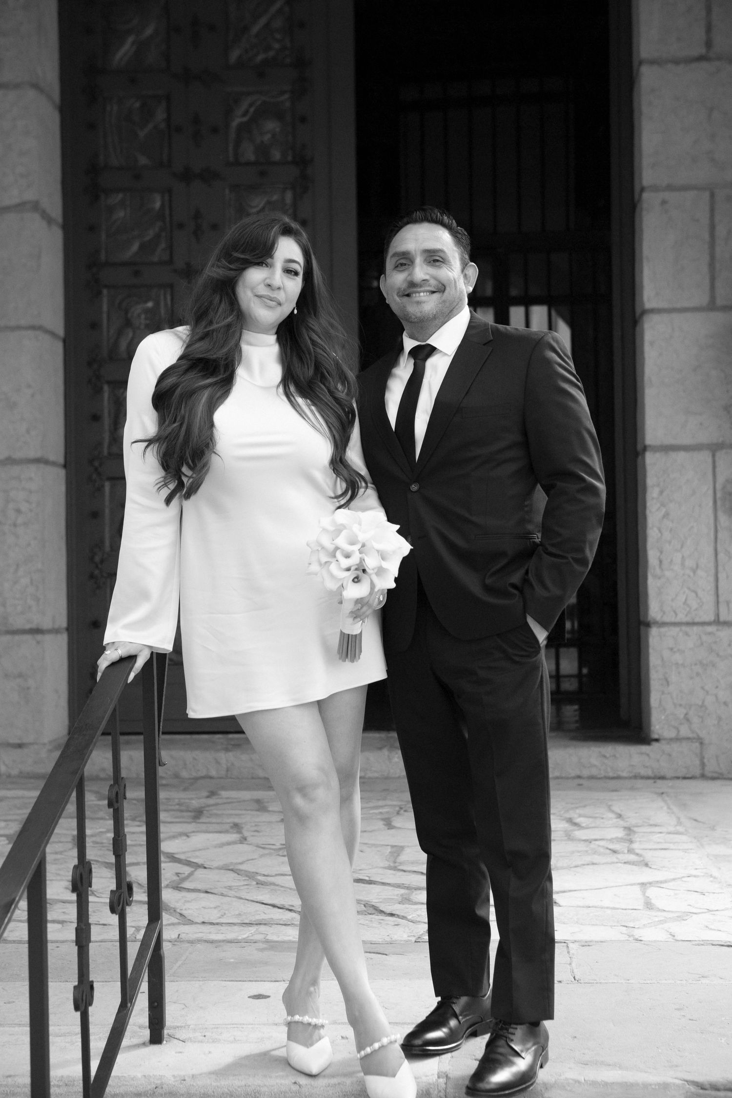 A bride in a short white dress and groom in black suit pose on stone steps in a black and white wedding photo.