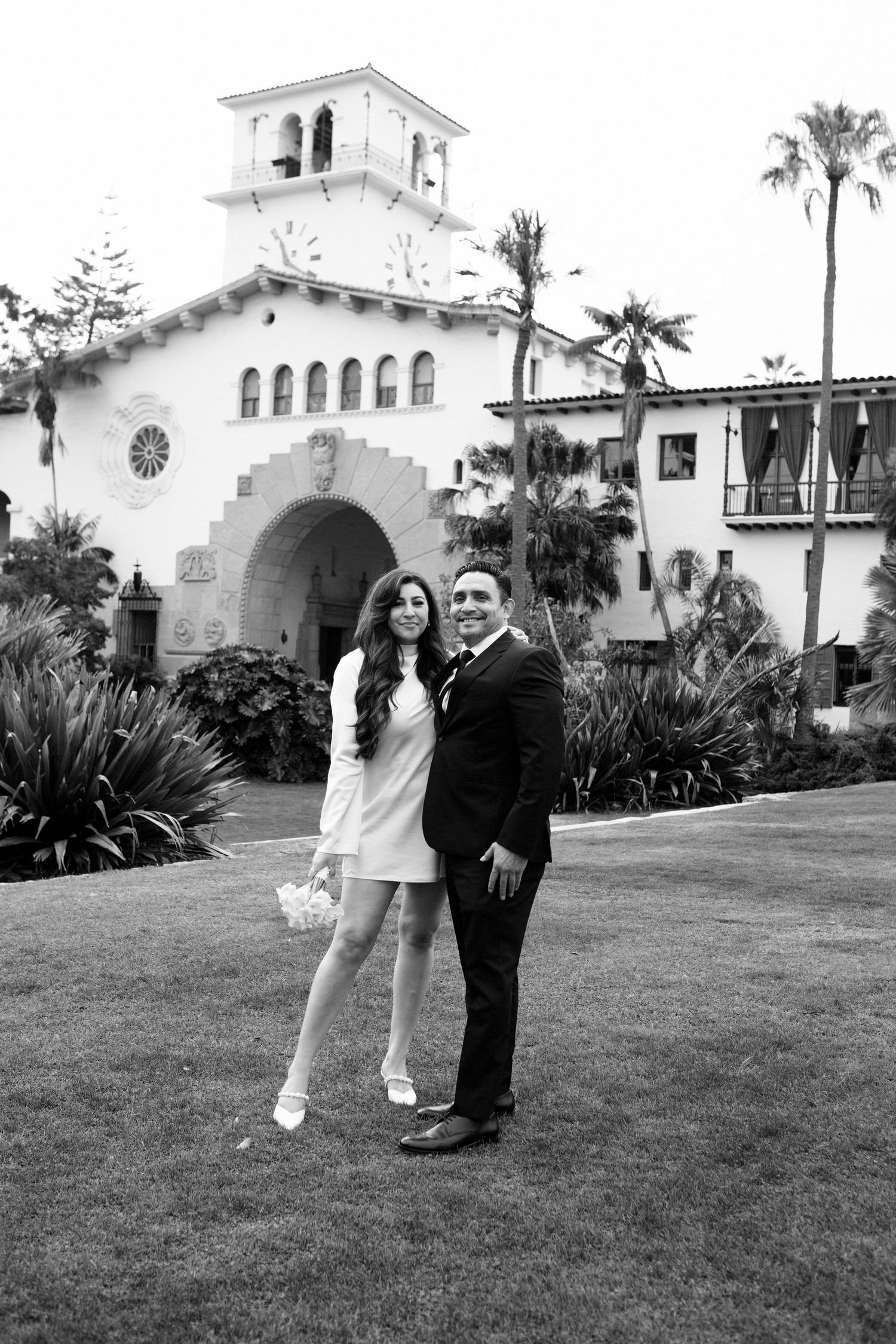 A couple poses on a lawn in front of a Spanish colonial style building with white stucco walls and arched doorways.
