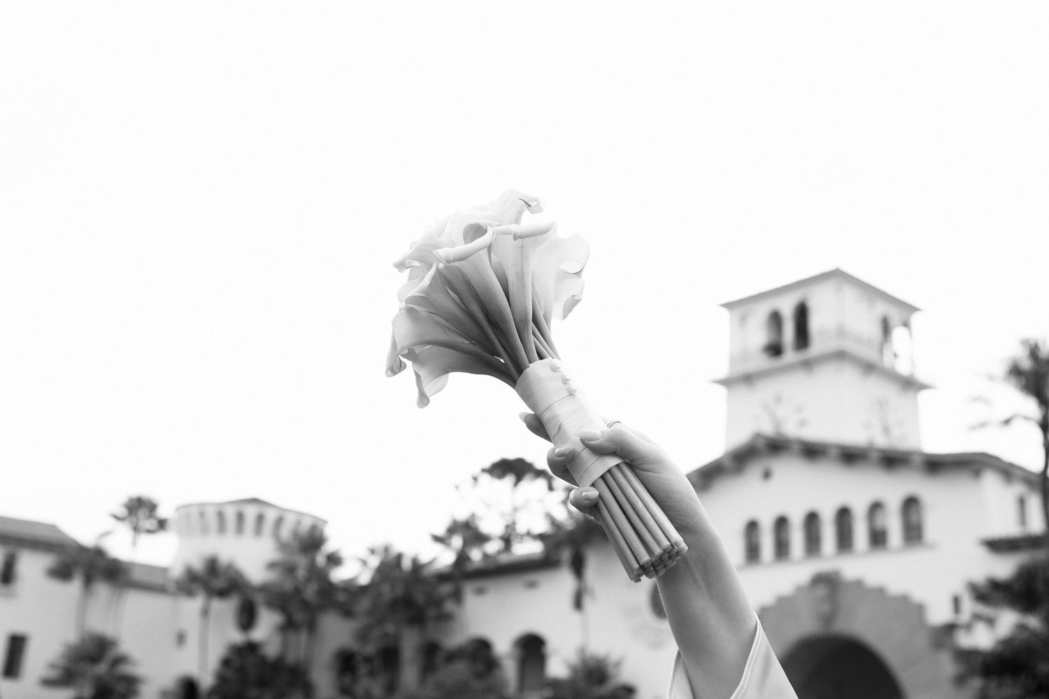A hand holds up a white bouquet against a Spanish-style building with a tower in black and white photography.