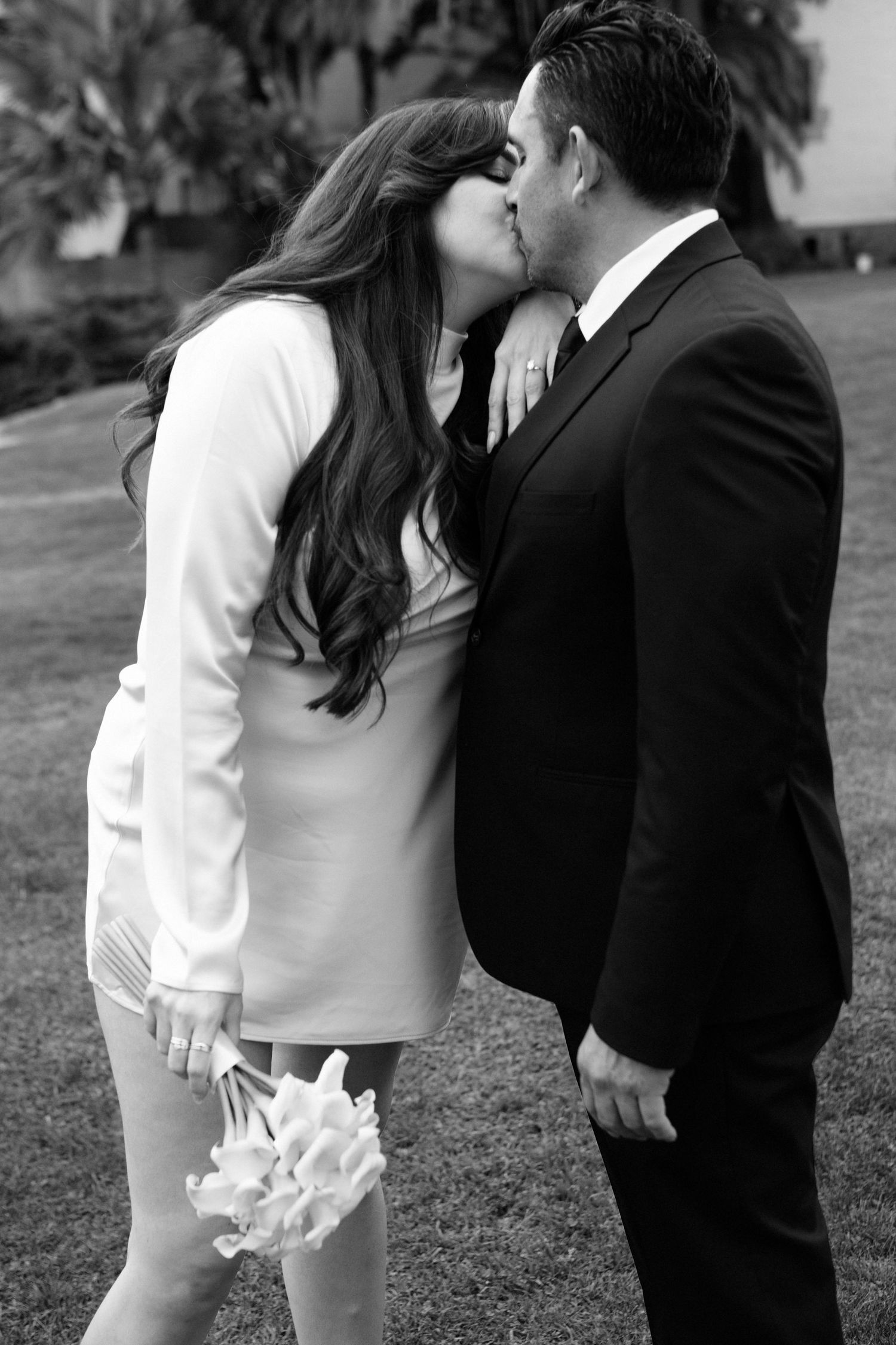 A romantic black and white wedding photo showing a couple sharing a kiss outdoors, with the bride holding a small bouquet.