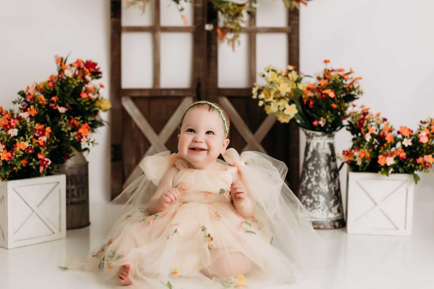 Baby in a pink tulle dress sits on floor surrounded by orange and yellow floral arrangements.