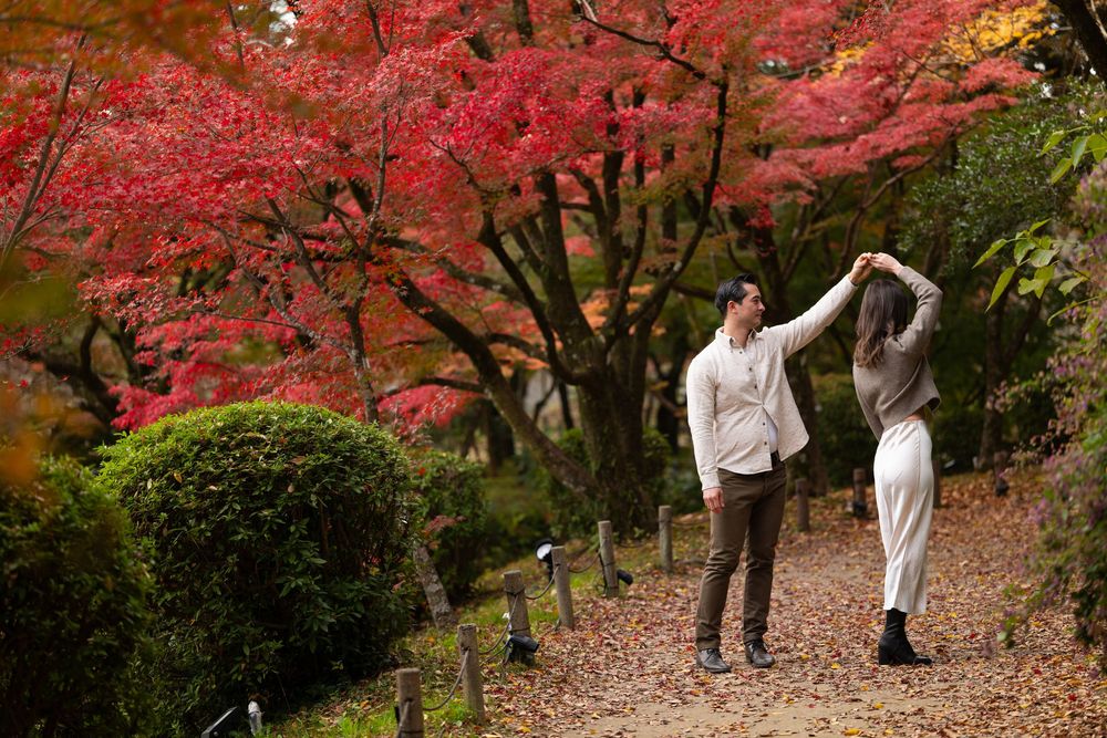 Man twirls woman under brilliant red maple trees on a scenic autumn garden path.