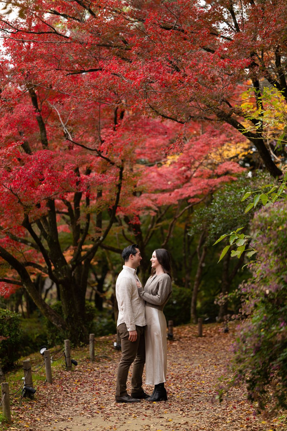 Couple embraces and smiles together on a leaf-strewn path beneath vibrant red and orange autumn maple trees.