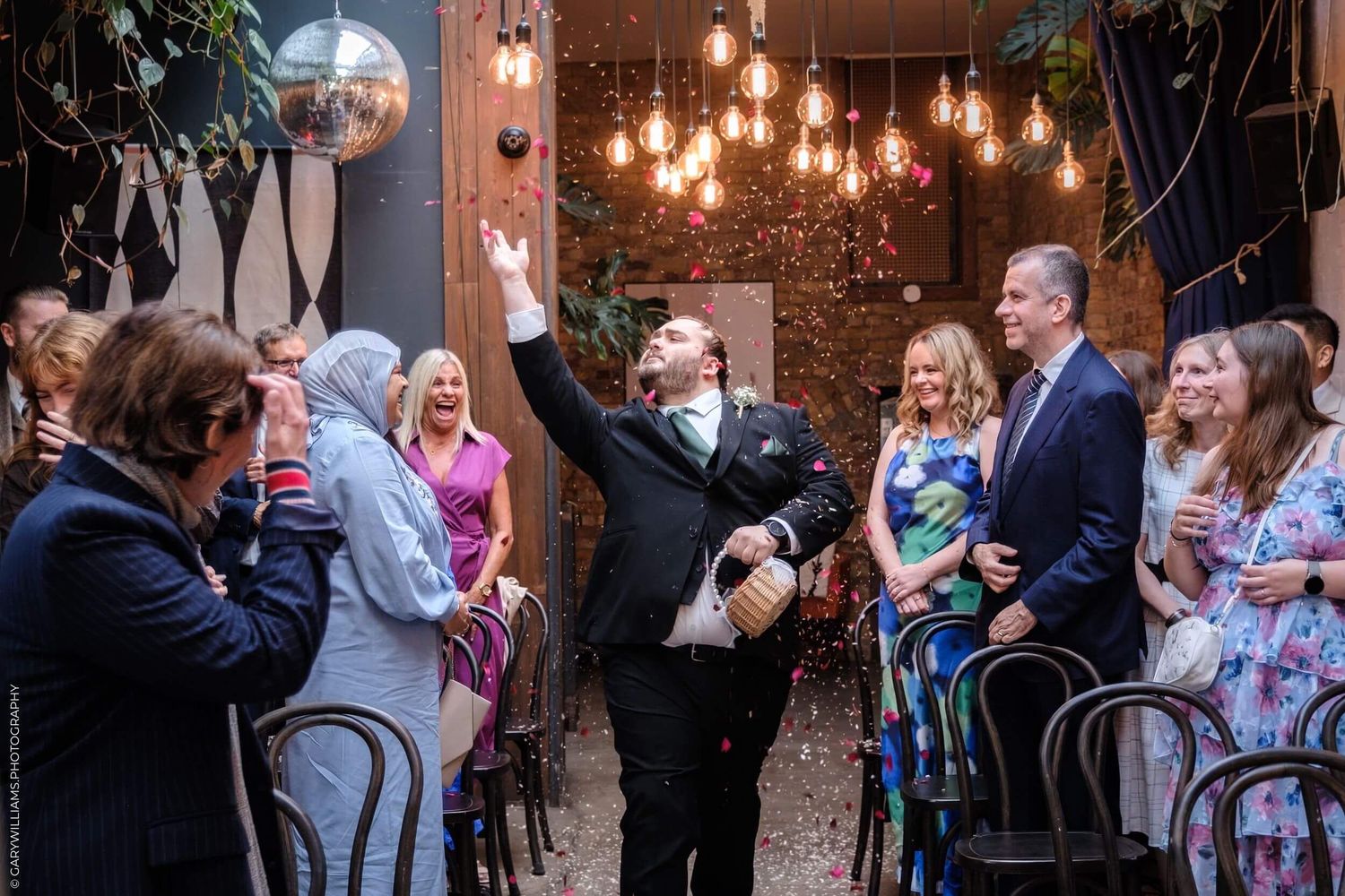 Wedding guests dance and celebrate during a reception in a rustic barn venue illuminated by warm hanging lights.